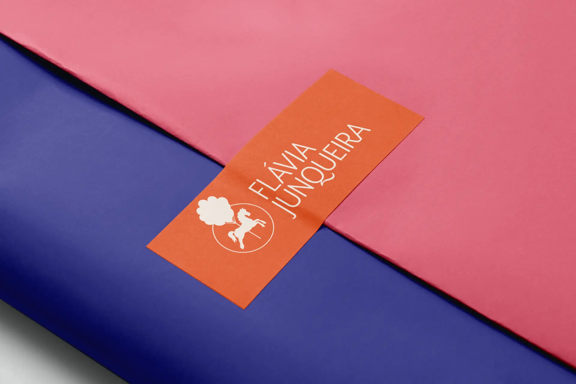Cartão laranja com a marca Flávia Junqueira sobre papéis rosa e azul